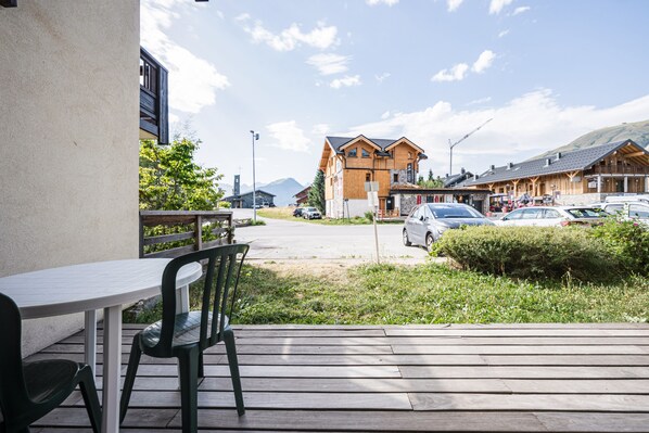 Une terrasse en bois avec une table et des chaises, donnant sur un parking où se trouvent des voitures et un bâtiment doté d’un toit en pente.