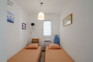 A small, clean room with two single beds, a window with blinds, a ceiling light, a wall-mounted heater, and a framed picture. - La Parenthèse de Pentrez - 100 m de la plage (Saint-Nic)