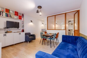 Un salon moderne avec un canapé bleu, une table à manger avec des chaises, un téléviseur et une bibliothèque remplie de livres et d’objets décoratifs.