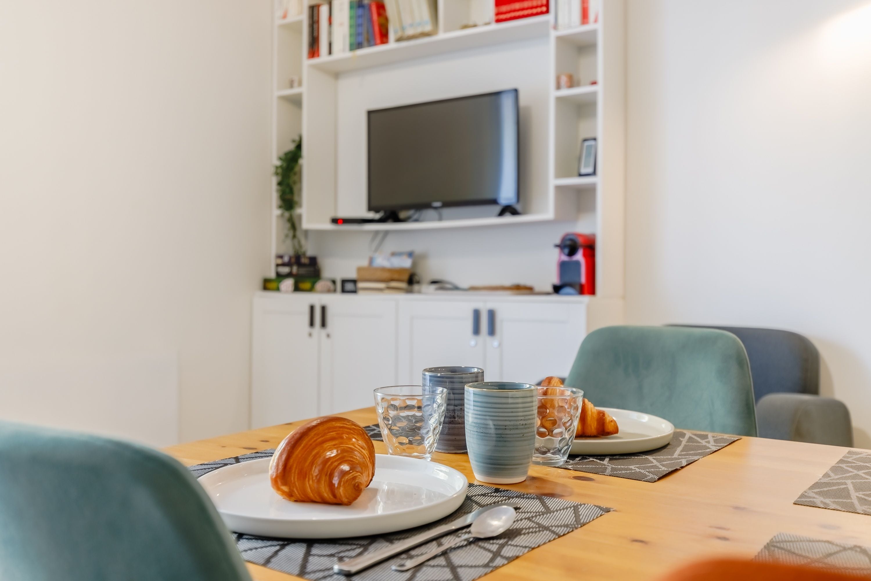 Un coin repas avec une table dressée pour deux personnes, un téléviseur fixé au mur blanc et une bibliothèque en arrière-plan.