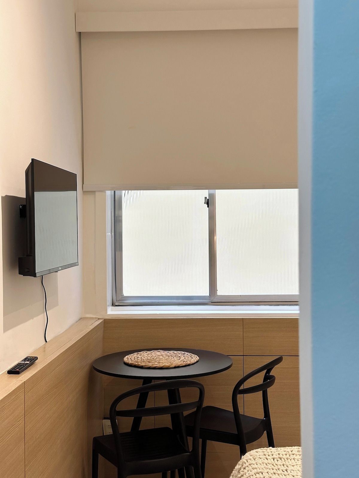 Una habitación pequeña con una televisión montada en la pared, una mesa redonda con sillas y una ventana con persianas.