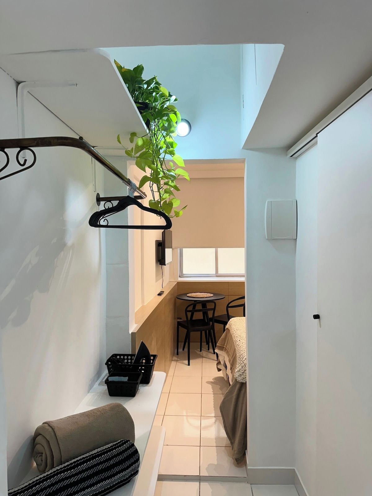 Una habitación compacta con cama, una mesita pequeña y sillas, una planta colgante y una toalla en el suelo.