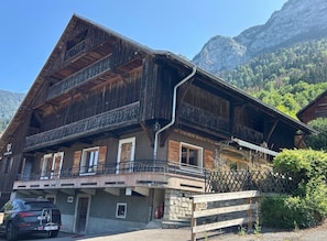 A wooden chalet with a balcony, stone foundation, and a mountain backdrop. - CHALET MONTAGE, a renovated former farmhouse located between the Arve Valley and the ski resorts (Glières-Val-de-Borne)