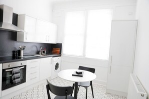 A compact kitchenette with a built-in oven, a washing machine, and a small dining table with chairs. - Spacious Ground-Floor Leith apartment | Sleeps 6 (Edinburgh)