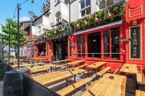 Ein Sitzbereich im Freien mit Holztischen und Bänken, eine rote Bar mit einem Schild, auf dem 'IRISH BAR' steht, und ein Gebäude mit einem 'GUINNESS'-Schild über dem Eingang.