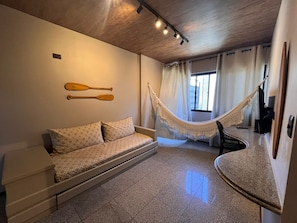 Ein Zimmer mit einer Couch, einer Hängematte, einem an der Wand befestigten Fernseher und einem Fenster mit Vorhängen.