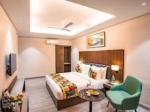 A hotel room with a bed, a ceiling fan, a painting, a nightstand, a chair, and a small table with a vase and fruit. - Classic King Room (Agra)