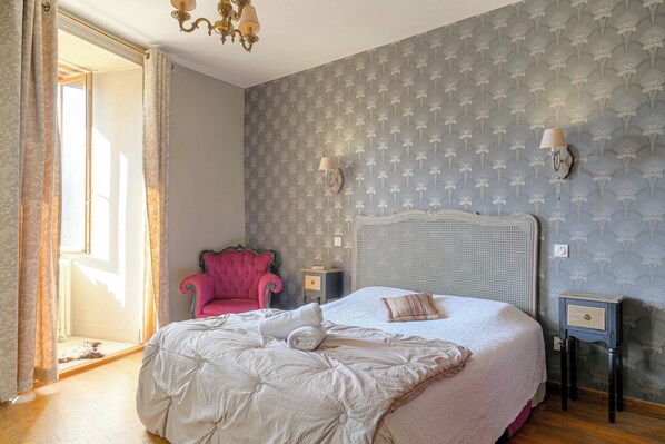 Een slaapkamer met een groot bed, een chaise longue, een kroonluchter en behang met een patroon.