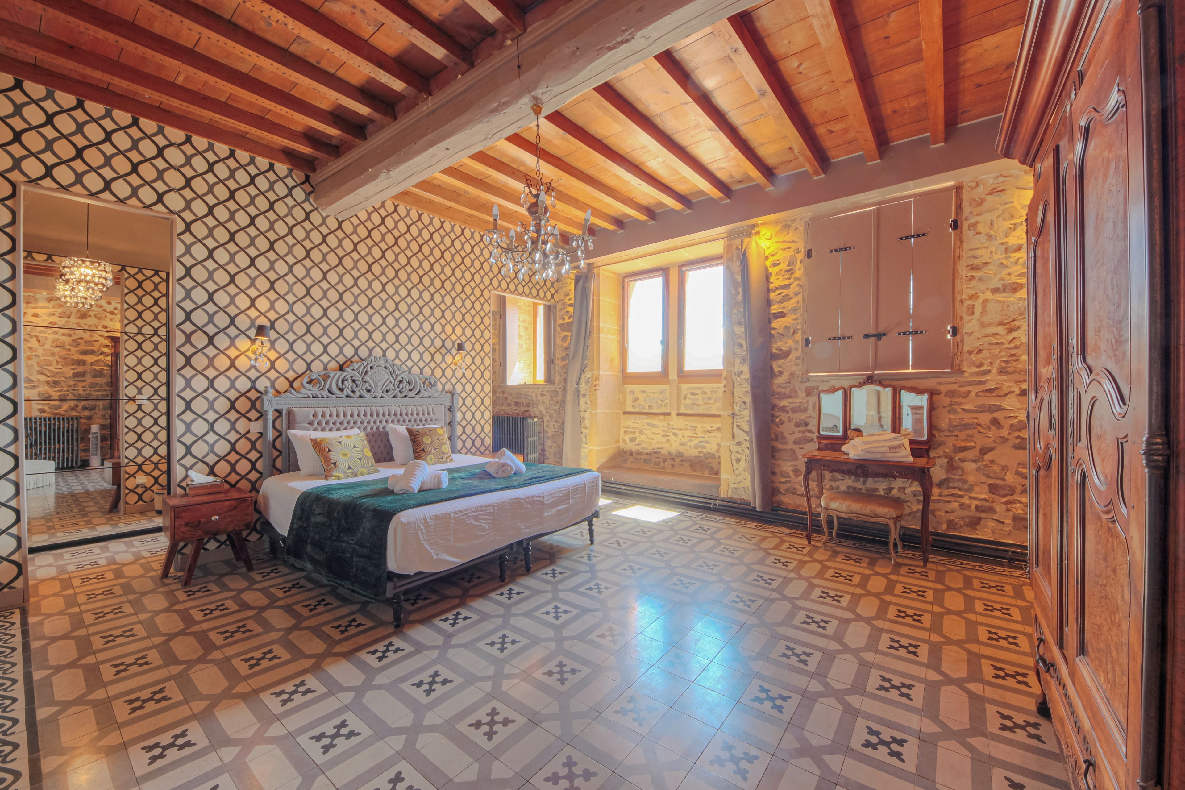 A bedroom with a large bed, a chandelier, a stone wall, and a wooden ceiling.