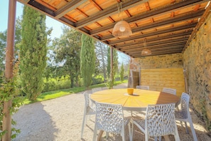 Un patio exterior con una mesa redonda de madera y sillas blancas, rodeado de grava y follaje.
