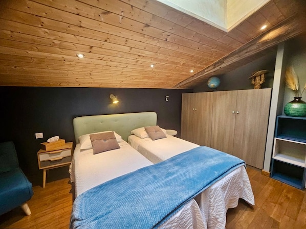 A bedroom with a wooden ceiling, two beds, a nightstand, a wardrobe, and a shelf with decorative items.