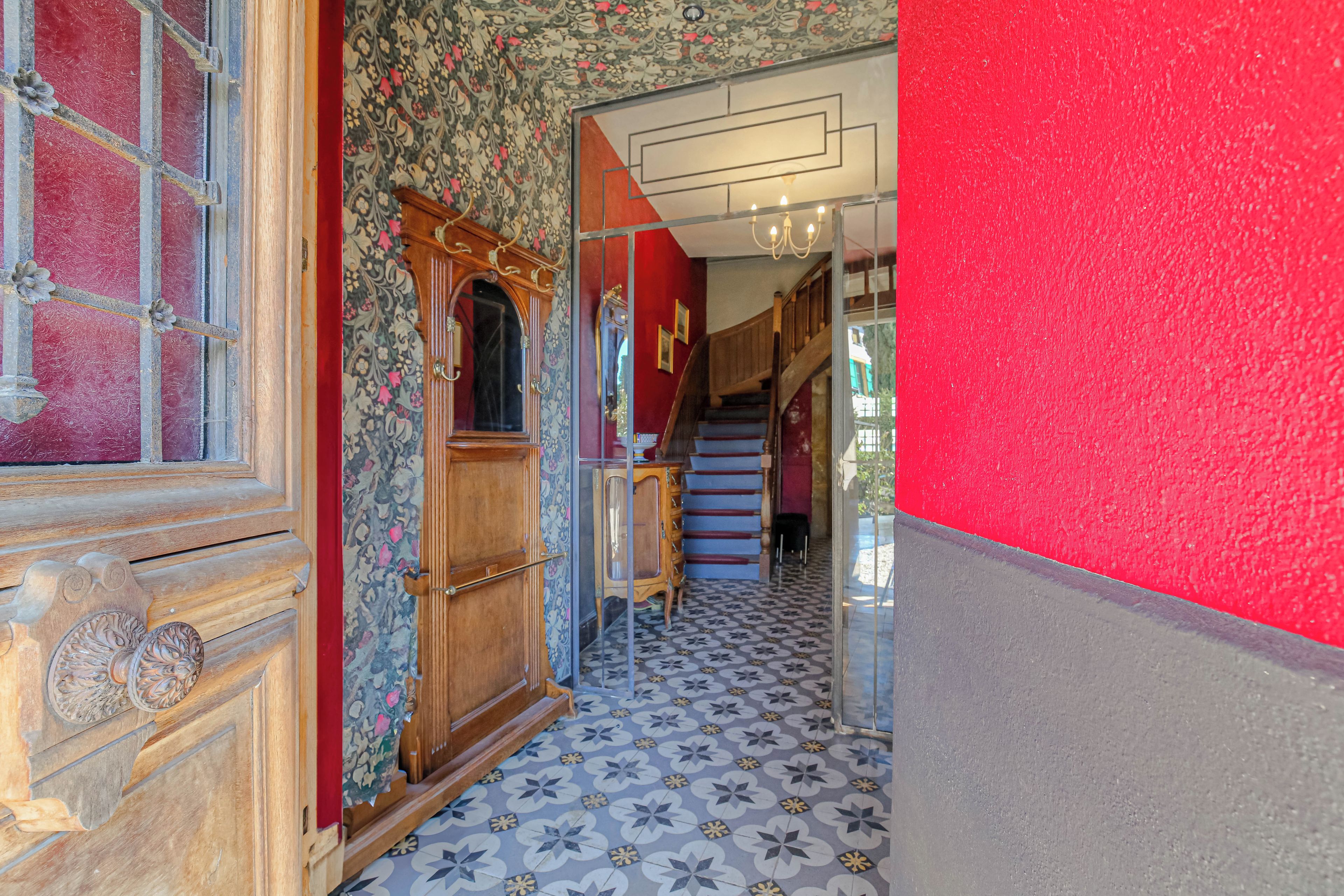 Un couloir avec des carreaux de sol à motifs, une porte en bois dotée d’un panneau de verre et un escalier avec un mur rouge d’un côté.