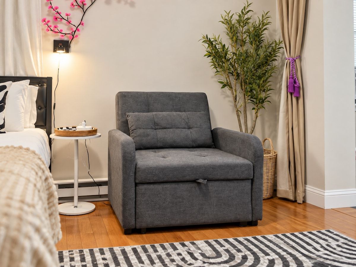 Un sillón gris en una habitación con una cama, una mesita con una bandeja, una planta en maceta y un tapete a rayas.