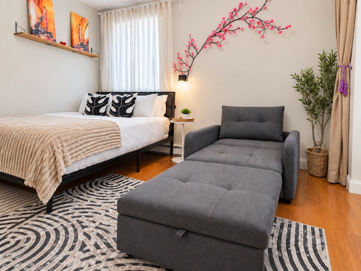 Une chambre à coucher moderne avec un lit, un canapé gris, une petite table d’appoint et une lampe fixée au mur.