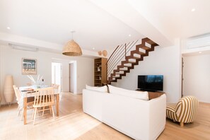 Ein modernes Wohnzimmer mit einem weißen Sofa, einem Essbereich mit Holztisch und -stühlen, einer außergewöhnlichen Treppe und einem Fernseher.