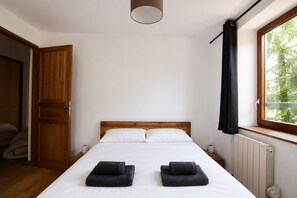 A bedroom with a wooden bed, white bedding, two rolled towels, a window with a view of trees, and a radiator. - NEW! Apartment Alpage | Large Terrace (Les Houches)