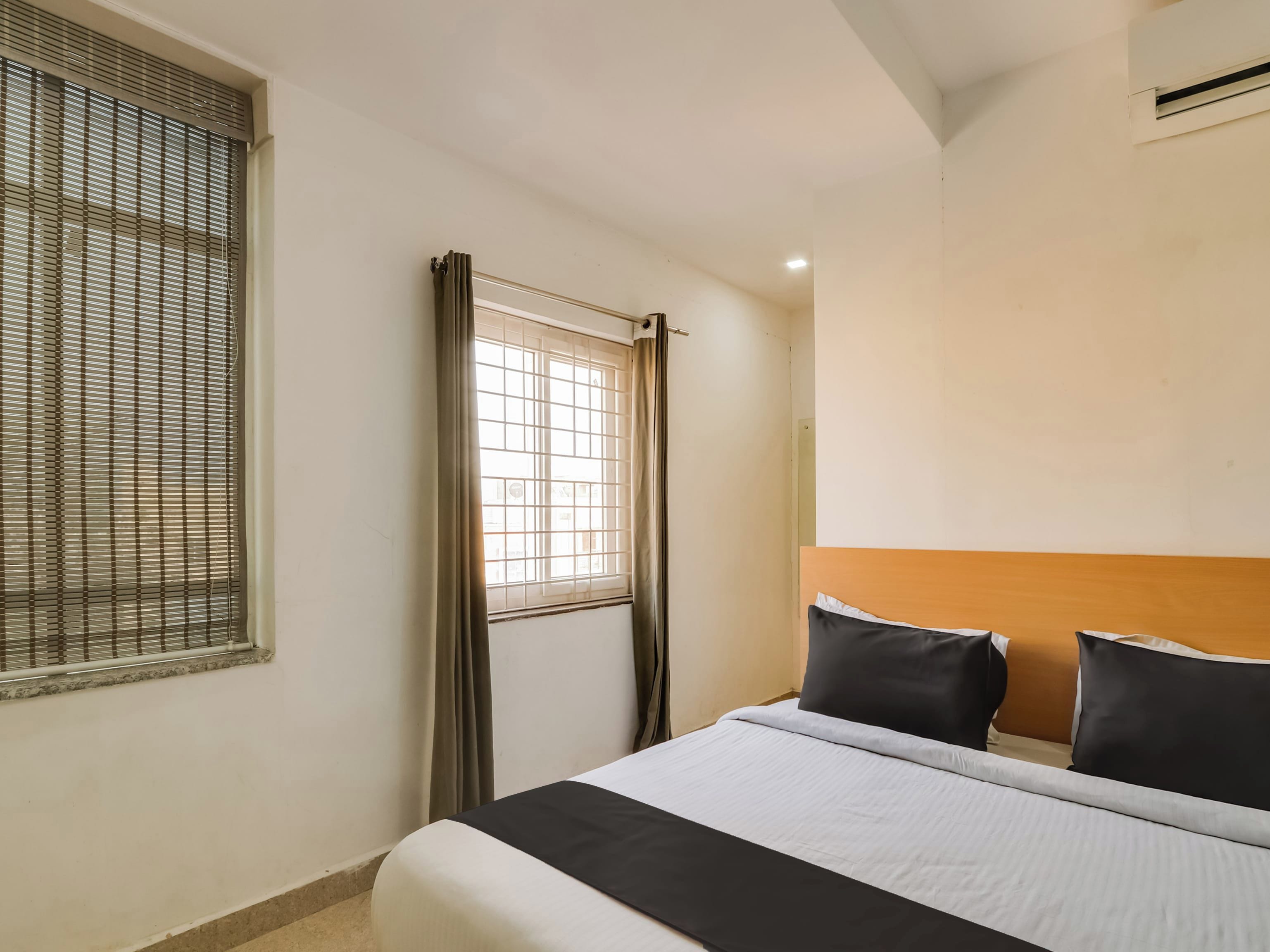 A hotel room with a bed, a window with blinds, a curtain, and an air conditioning unit.