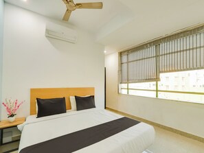 Habitación de hotel con cama, mesita de noche, ventilador de techo, aire acondicionado y ventana con persianas.