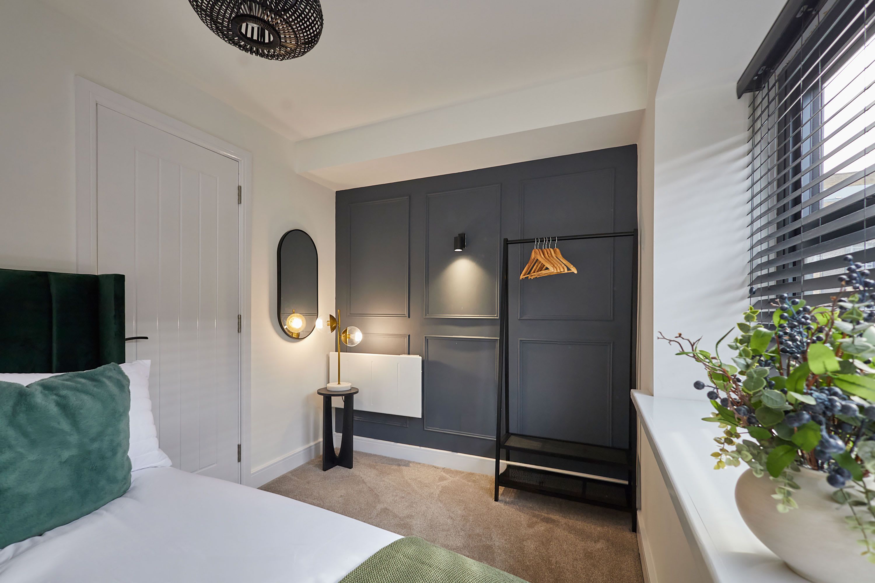 Quarto de hotel com cama, mesa de cabeceira, espelho, luminária pendente, radiador, cabideiro e janela com persianas.