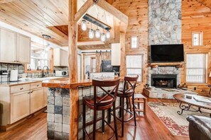 A log cabin interior with a stone fireplace, wooden bar, and kitchen area. - Wooded Wonder | Creek + Hot Tub + Game Room (Cleveland)