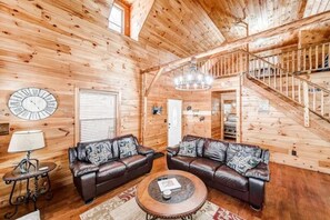 Ein Holzblockhaus-Innenraum mit einem Wohnzimmer, ausgestattet mit zwei Ledersofas, einem runden Couchtisch, einem Kronleuchter und einer Treppe.