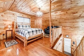 A wooden cabin bedroom with a bed, a nightstand, a lamp, a window with blinds, a ceiling light fixture, and a door. - Wooded Wonder | Creek + Hot Tub + Game Room (Cleveland)