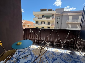 Un balcon avec une table et des chaises bleues, un objet jaune sur la table et un sol à motifs.
