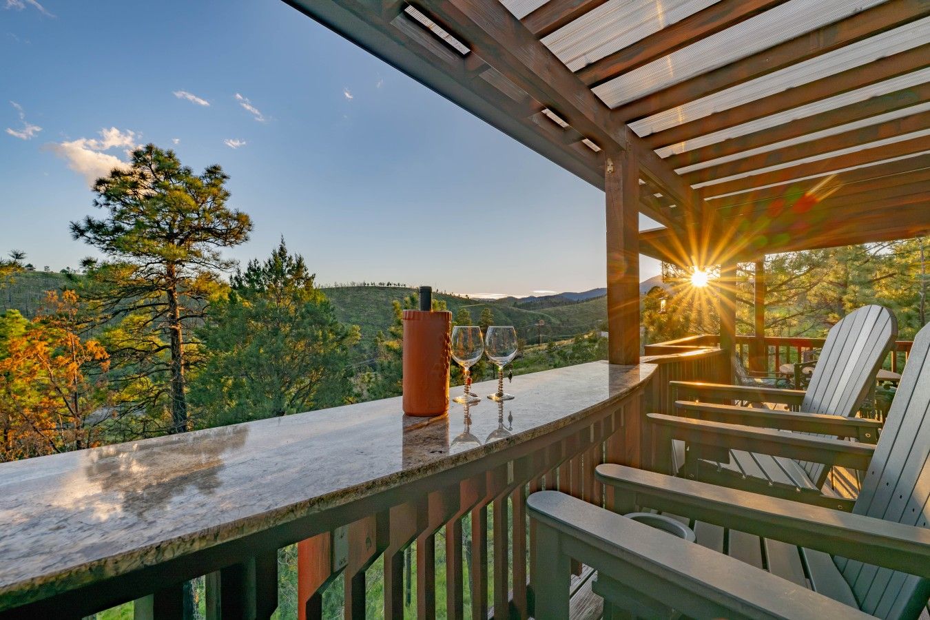 A wooden deck with Adirondack chairs, a bottle, and two wine glasses, overlooking a mountainous landscape.