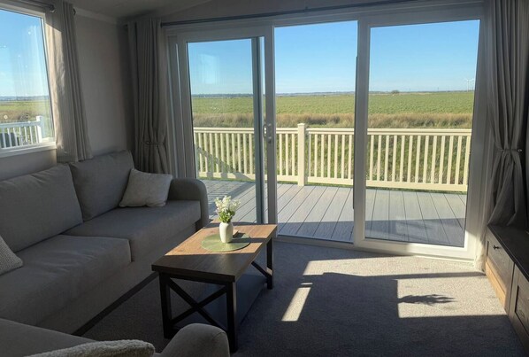A room with a sofa, a small table with a vase, and a view of a deck and grassy field through glass doors.