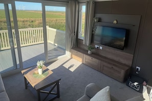 A room with a TV, a wooden table with a plant, a sofa, and a view of wind turbines.