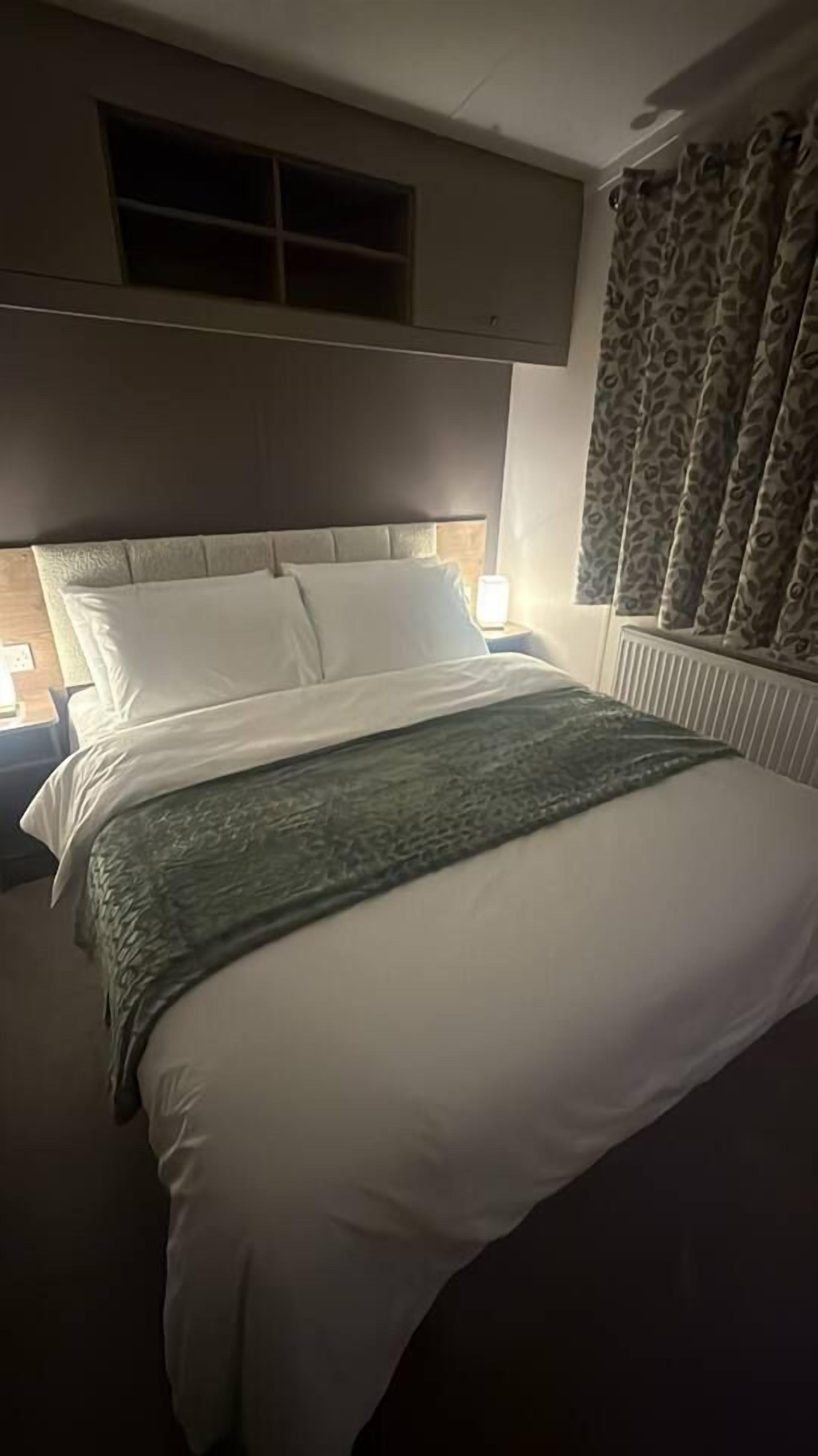 A hotel room with a bed, a nightstand, a window with curtains, and a radiator.