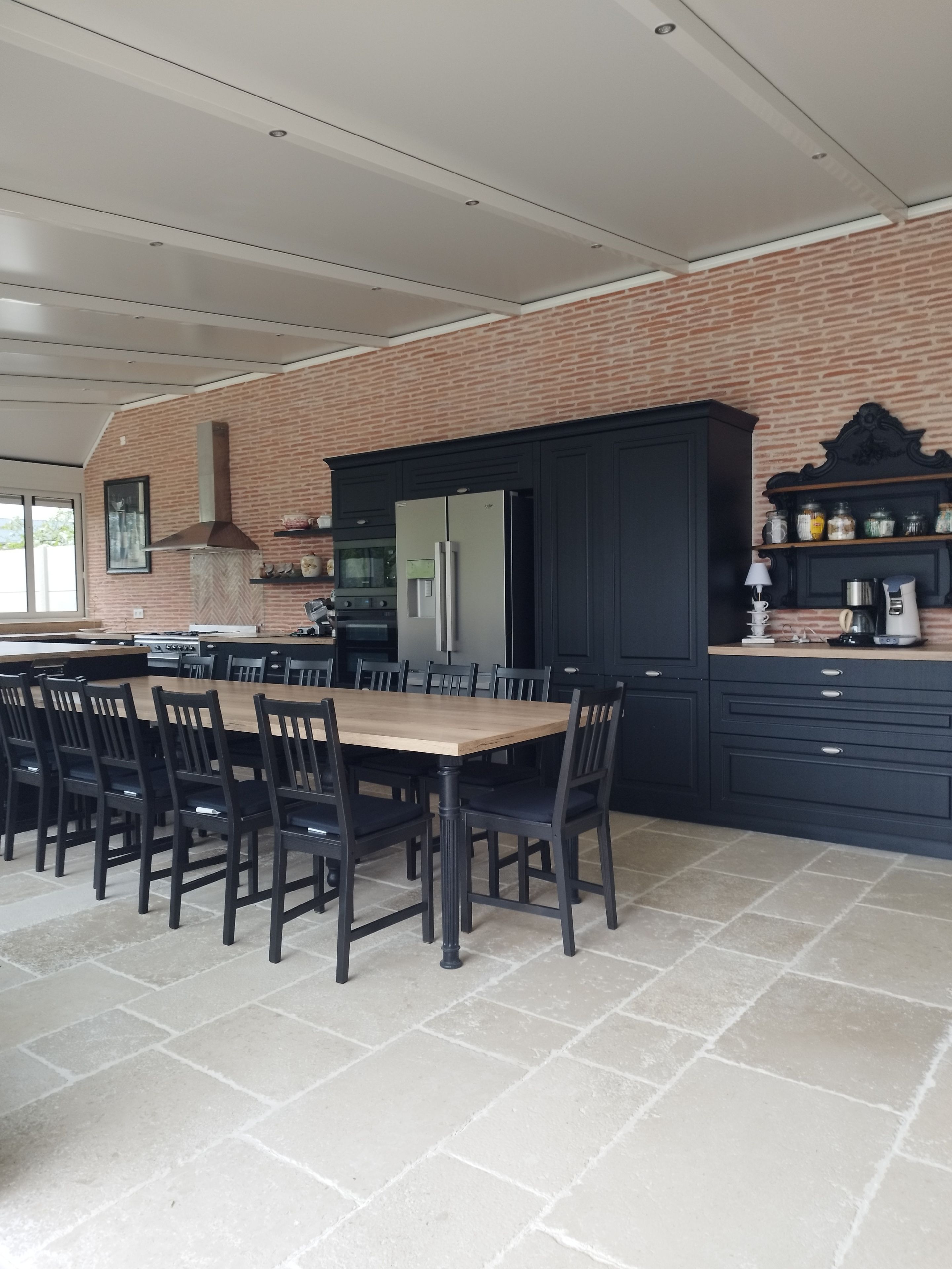 A kitchen with a brick wall, dark cabinetry, a stainless steel refrigerator, and a dining table with chairs.