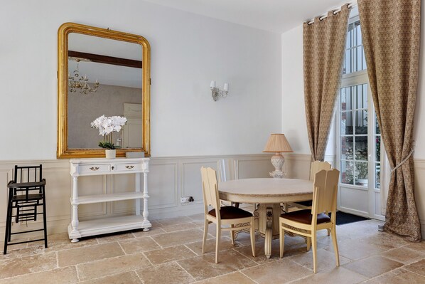 A room with a round table, two chairs, a white console with a mirror, a lamp, and a window with curtains.