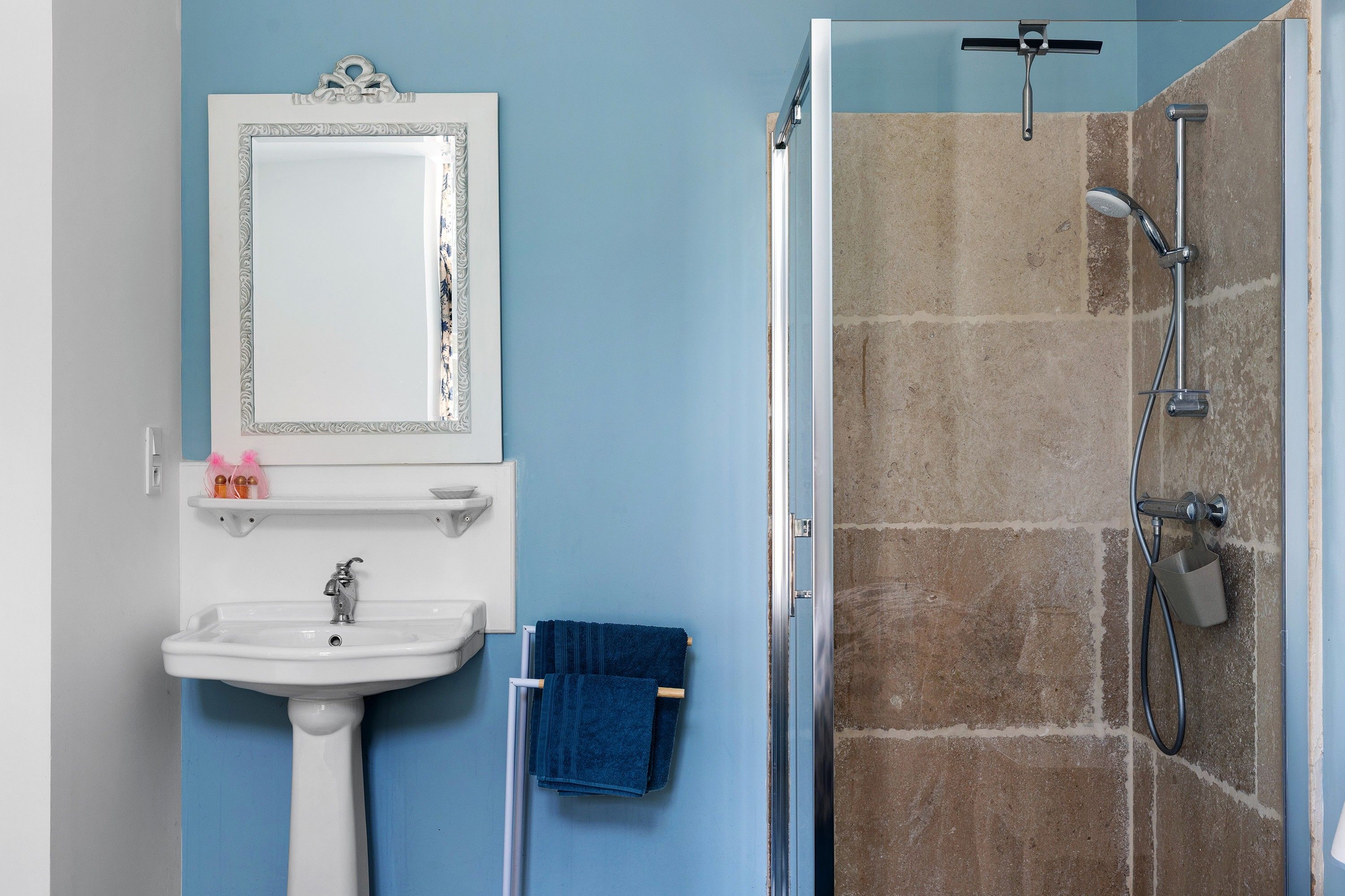 Un baño con lavamanos blanco, un espejo con marco decorativo, una toalla azul y una zona de ducha con pared de azulejos.