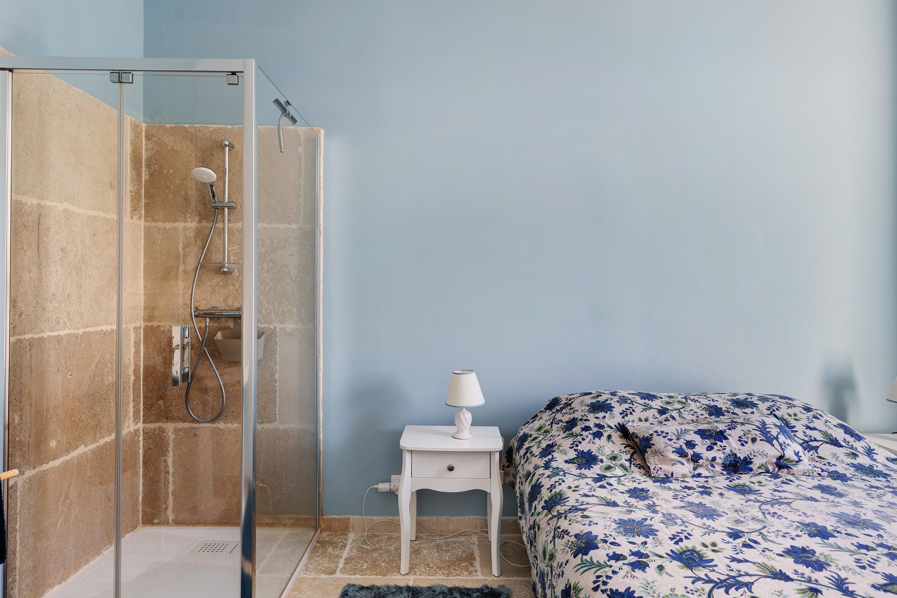 Une salle de bain avec une douche à parois de verre, une table de chevet blanche avec une lampe et un lit recouvert d’une couvre-lit à motifs floraux.