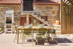 Un área de comedor al aire libre con un muro de piedra de fondo, una mesa de madera dispuesta para cuatro personas y sillas de metal verde.