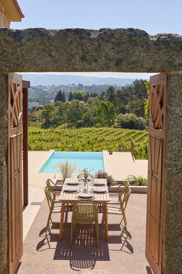 Un área de comedor al aire libre con piscina, rodeada por un muro de piedra y vistas a un viñedo.