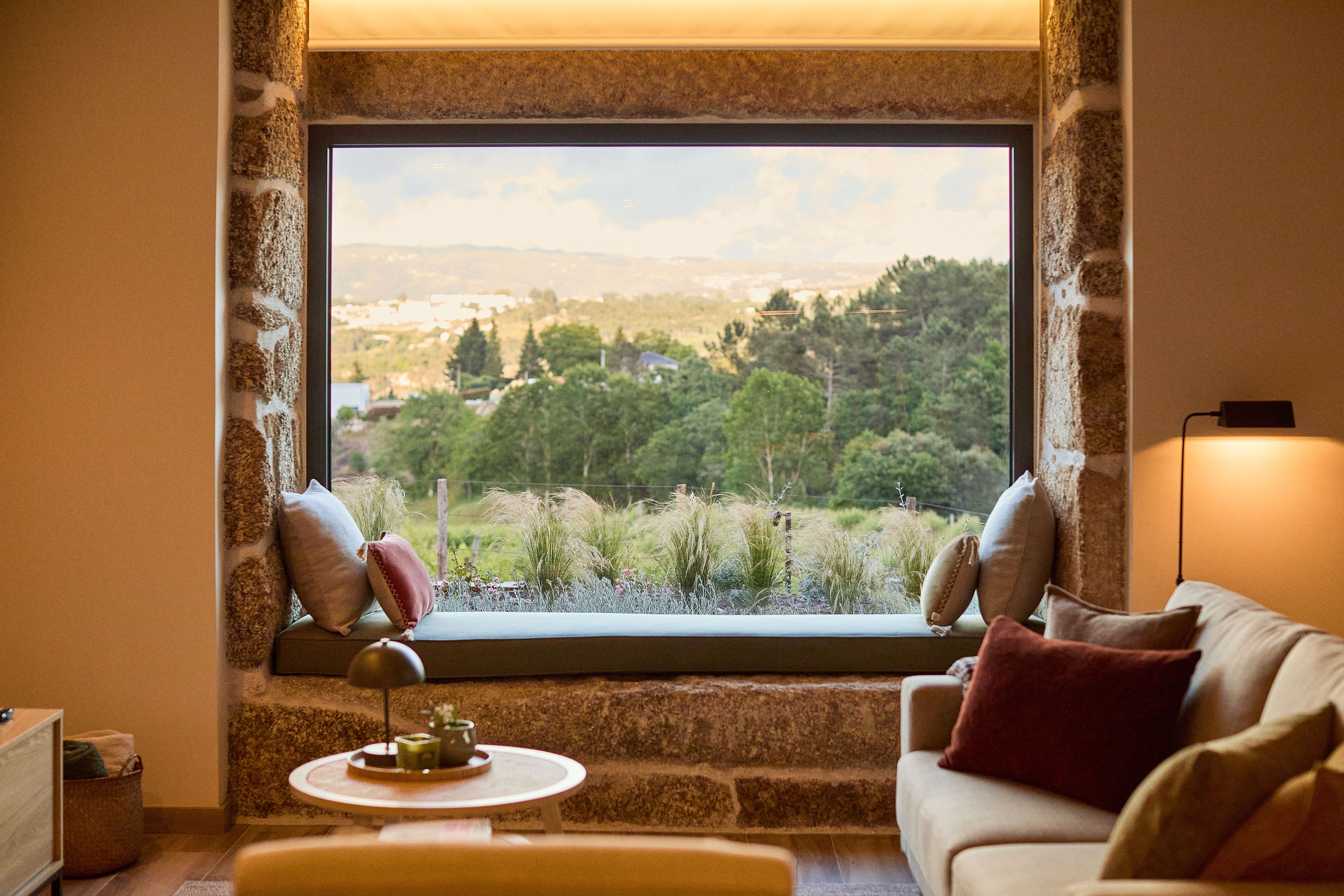 Une pièce chaleureuse avec un mur en pierre, une fenêtre donnant sur un paysage verdoyant, un canapé avec des coussins et une petite table avec une lampe.