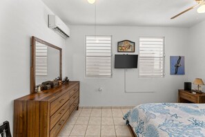 A bedroom with a wooden dresser, a bed with a blue patterned bedspread, a flat-screen TV mounted on the wall, and a ceiling fan. - Casa Pueblo Mar: Rincón Town Living with Sunset Sea Views (Rincon)
