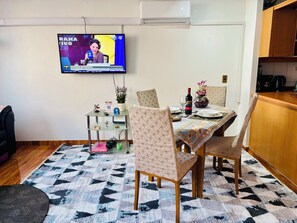 A dining area with a television displaying a news program, a table set with plates, glasses, and a bottle, and chairs with a patterned tablecloth. - Great Space for Remote Work, 5 Minutes from the Park (Santiago)