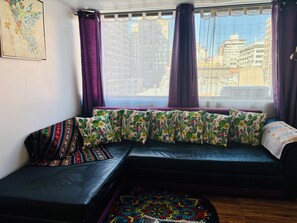 A room with a black leather sofa, a colorful patterned cushion, a window with purple curtains, and a cityscape view. - Great Space for Remote Work, 5 Minutes from the Park (Santiago)
