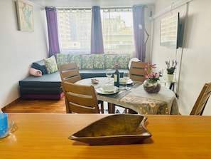 A dining area with a table set for two, a sofa with patterned cushions, and a view of buildings through the window. - Great Space for Remote Work, 5 Minutes from the Park (Santiago)