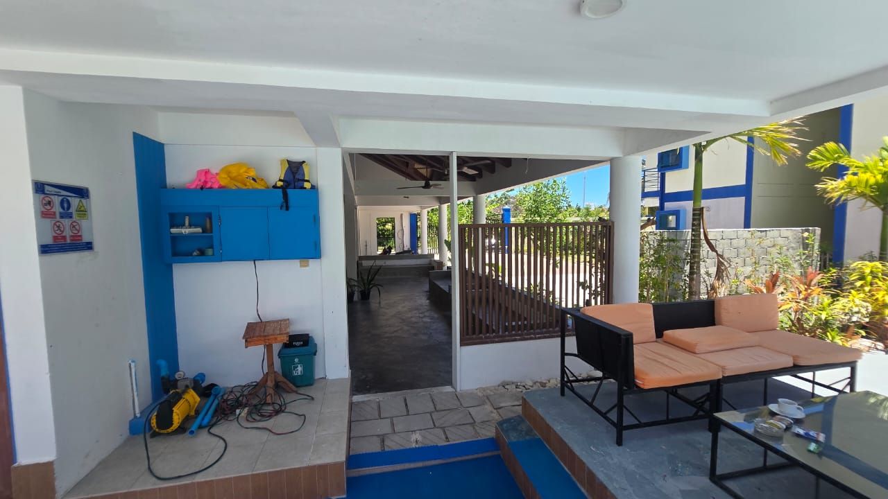 A covered outdoor seating area with a table and chairs, a blue storage unit, and a pathway leading to a building entrance.