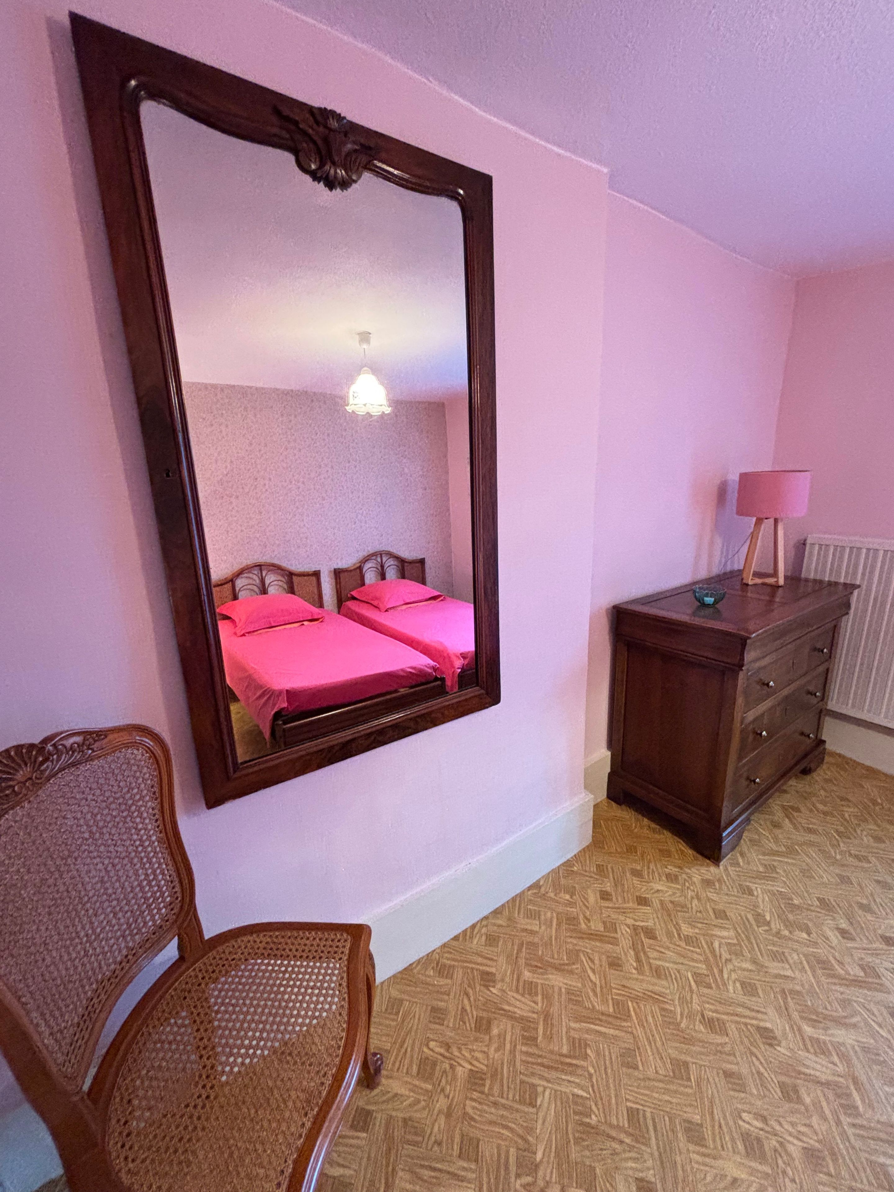 Une pièce avec un grand miroir, un lit avec une literie rose, une commode en bois et une chaise en osier.