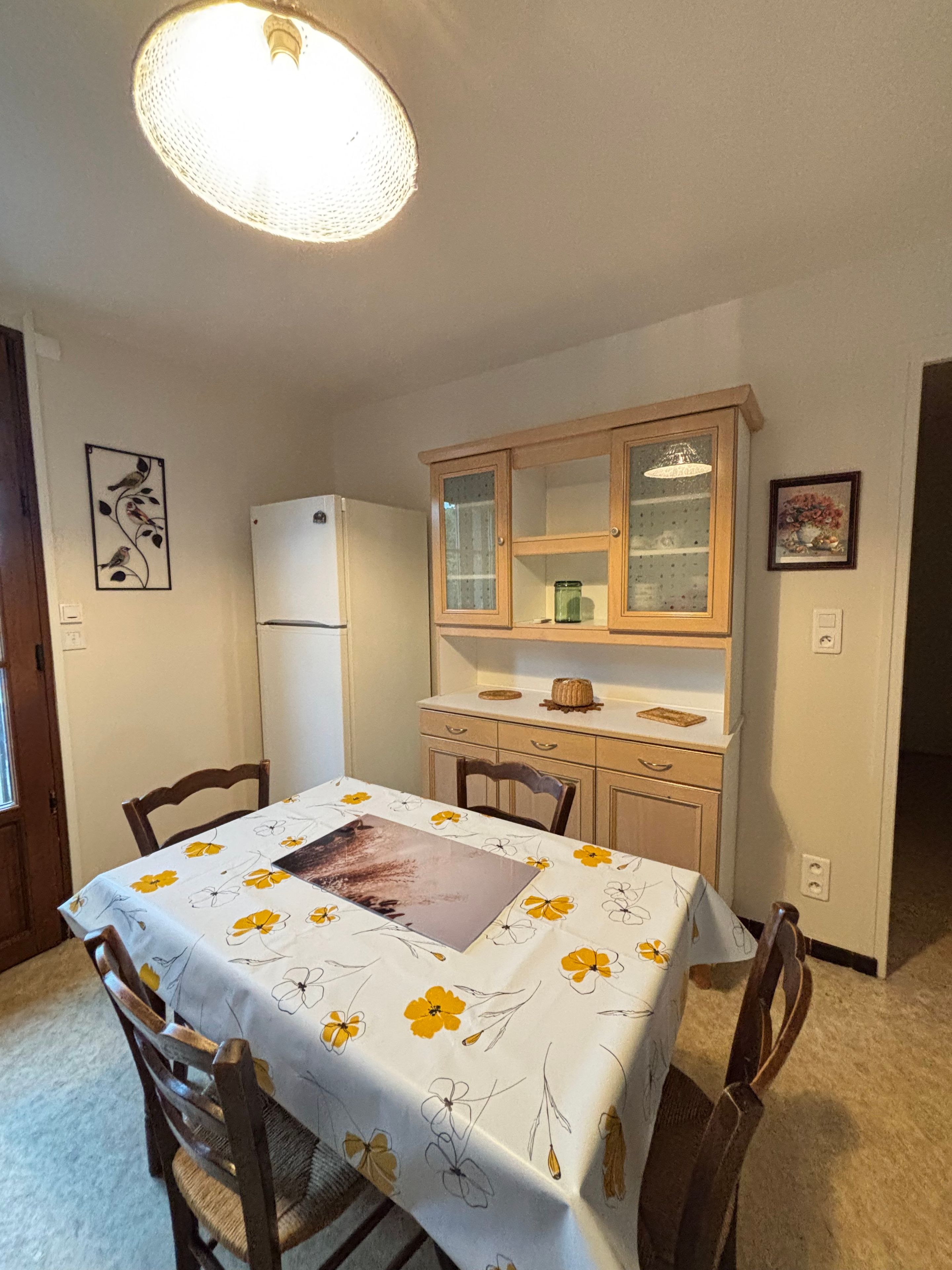 Un coin repas avec une table dressée pour quatre personnes, un réfrigérateur, un meuble en bois et un luminaire fixé au plafond.