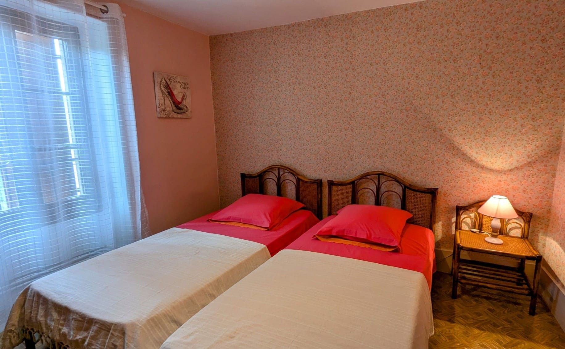 Une chambre avec deux lits, une table de chevet avec une lampe, une fenêtre avec des stores et un papier peint à motifs floraux.