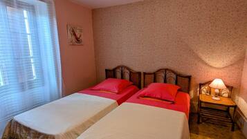 Une chambre avec deux lits, une table de chevet avec une lampe, une fenêtre avec des stores et un papier peint à motifs floraux.