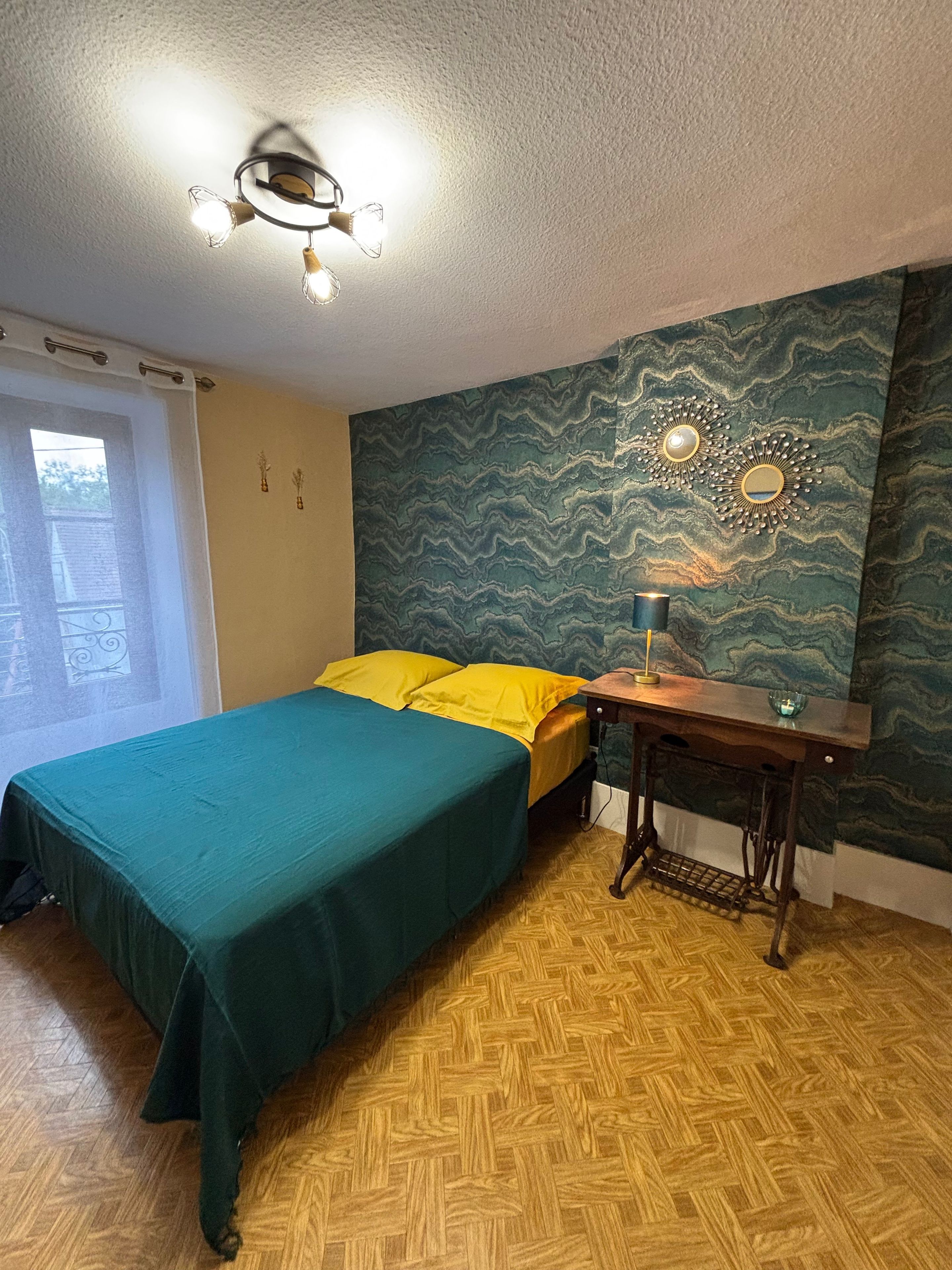 A room with a bed, a table, a chair, a window, and a wall with a decorative pattern.