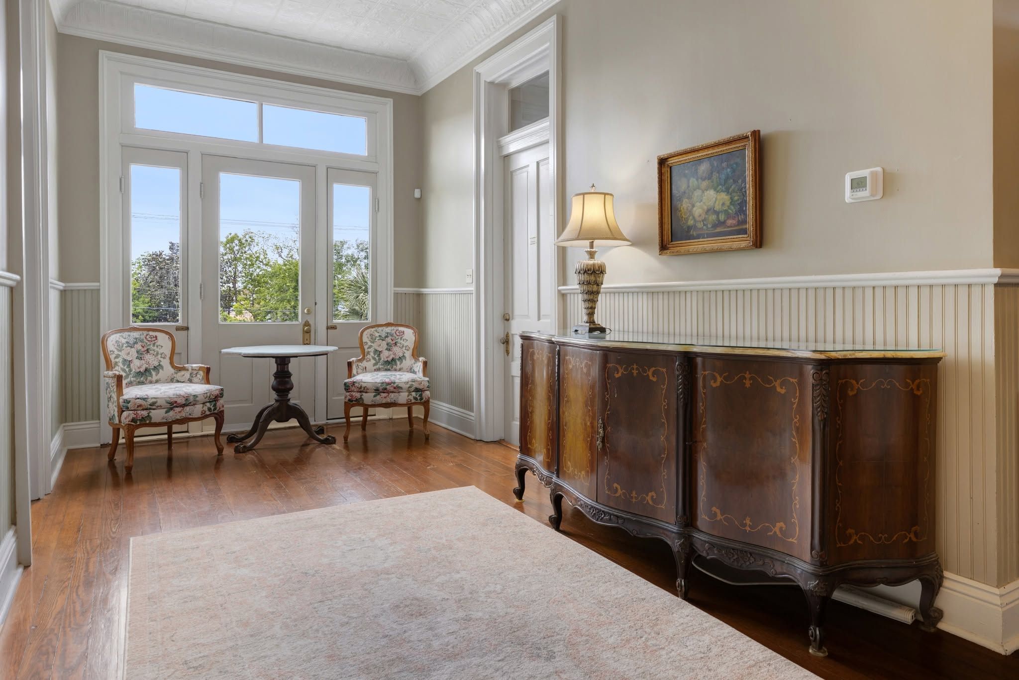 A room with a large window, a wooden cabinet, two chairs, a small table, and a lamp.