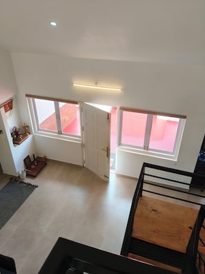 Una habitación con techo alto, dos ventanas, una puerta, un pequeño altar y una mesa.